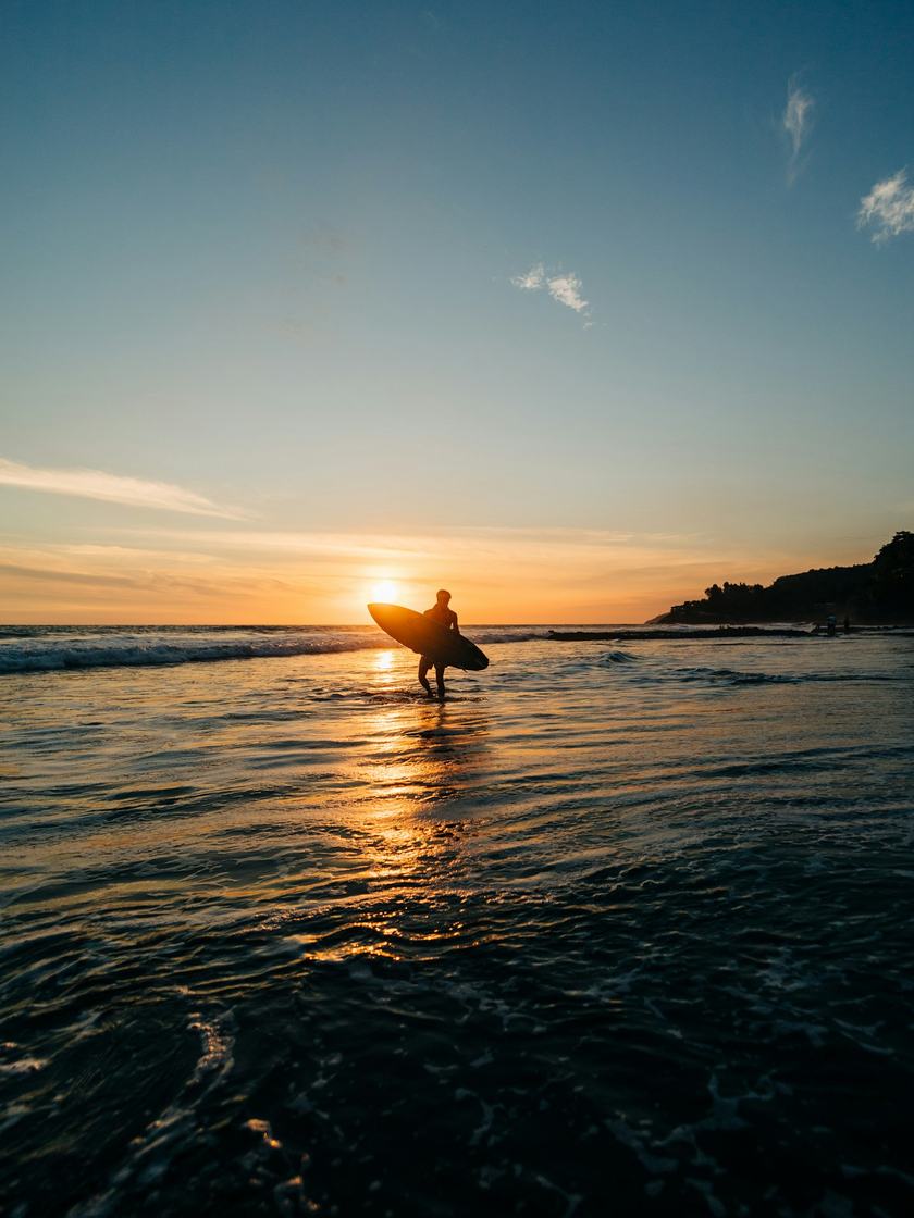Silueta de un surfista caminando por la orilla contra el sol que baja