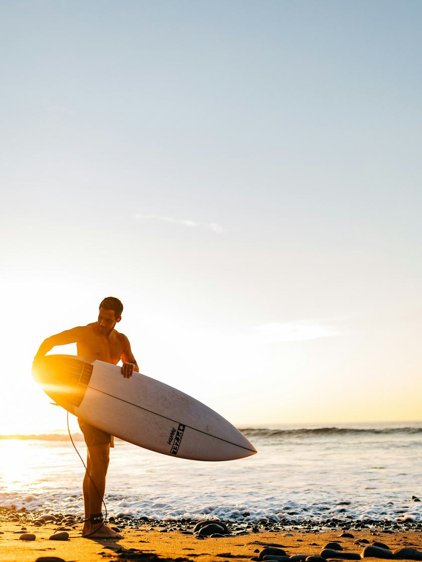 Un surfista sale del agua con la tabla bajo el brazo, a contraluz